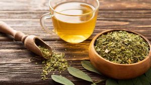 Cup of moringa tea beside a bowl of dried moringa leaves on a wooden table.