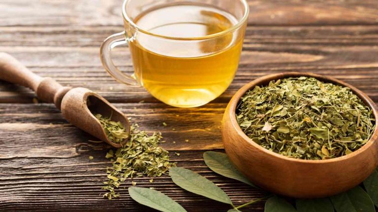 Cup of moringa tea beside a bowl of dried moringa leaves on a wooden table.