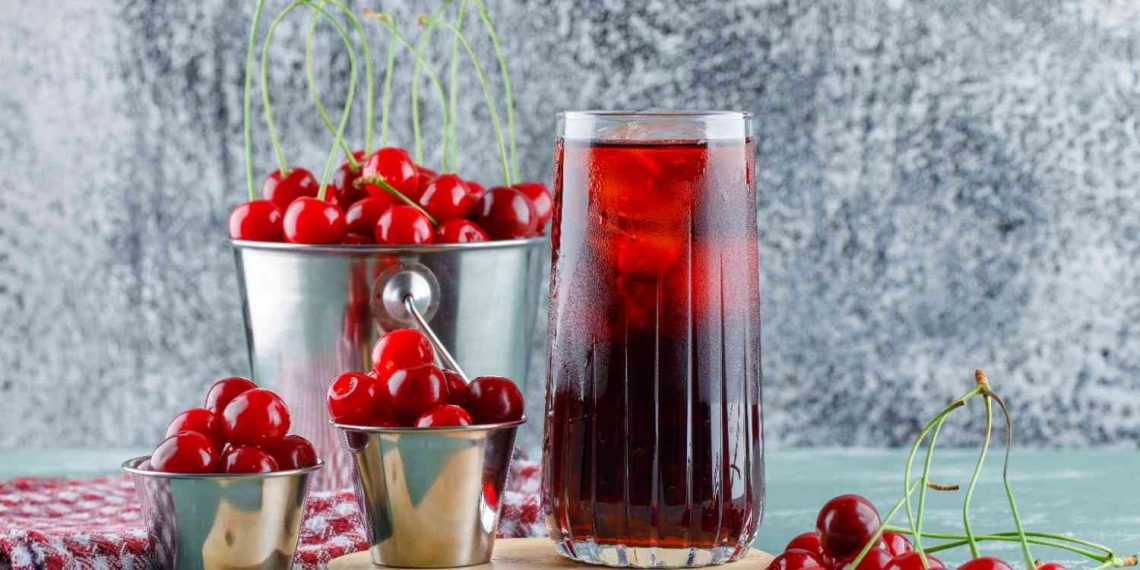 Cherry juice in a glass with ice next to fresh cherries in metal containers.