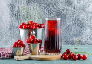 Cherry juice in a glass with ice next to fresh cherries in metal containers.