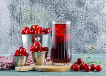 Cherry juice in a glass with ice next to fresh cherries in metal containers.