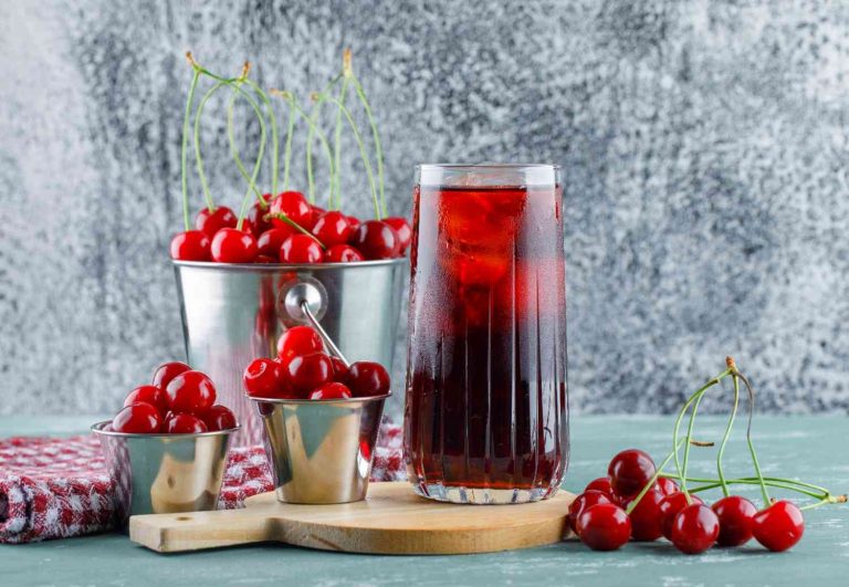 Cherry juice in a glass with ice next to fresh cherries in metal containers.