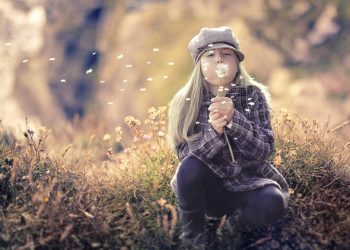 Girl in hat blowing dandelion seeds in a field.