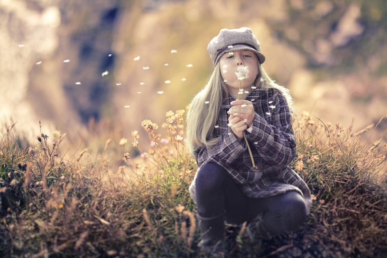 Girl in hat blowing dandelion seeds in a field.