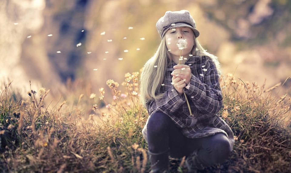 Girl in hat blowing dandelion seeds in a field.
