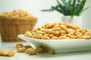 Salted peanuts piled on a white dish with a blurred basket and plant in the background.