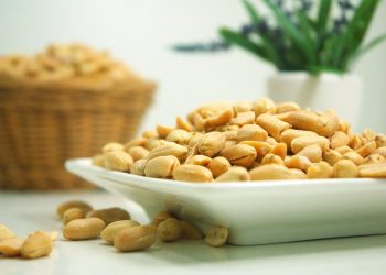 Salted peanuts piled on a white dish with a blurred basket and plant in the background.