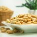 Salted peanuts piled on a white dish with a blurred basket and plant in the background.