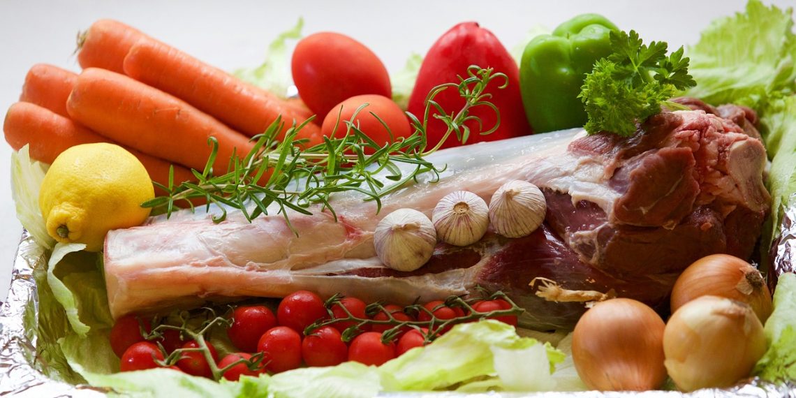 Roast lamb surrounded by fresh vegetables and herbs.