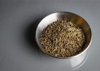 Bowl filled with cumin seeds on a gray surface.