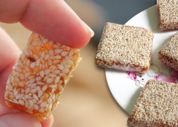Sesame candy square held by fingers next to plate of sweets.