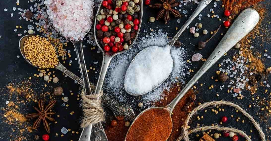 Various spices on spoons, including salt, pepper, mustard, and paprika.