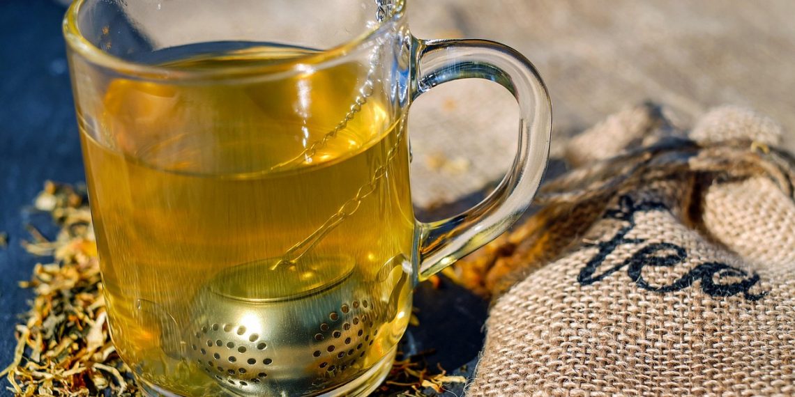 Glass of herbal tea with infuser next to burlap tea bag.