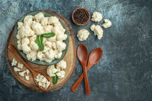 Cauliflower florets in a bowl with wooden spoons and peppercorns on a rustic surface.