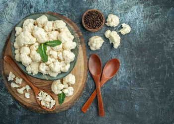 Cauliflower florets in a bowl with wooden spoons and peppercorns on a rustic surface.