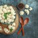 Cauliflower florets in a bowl with wooden spoons and peppercorns on a rustic surface.