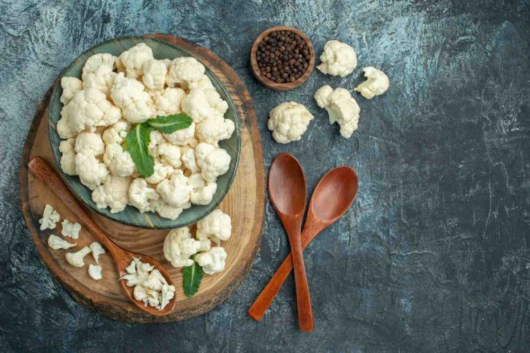 Cauliflower florets in a bowl with wooden spoons and peppercorns on a rustic surface.