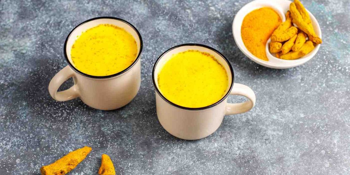 Two mugs of golden turmeric milk on a table with turmeric root and powder.