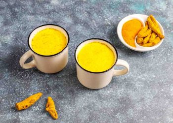 Two mugs of golden turmeric milk on a table with turmeric root and powder.