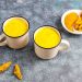 Two mugs of golden turmeric milk on a table with turmeric root and powder.