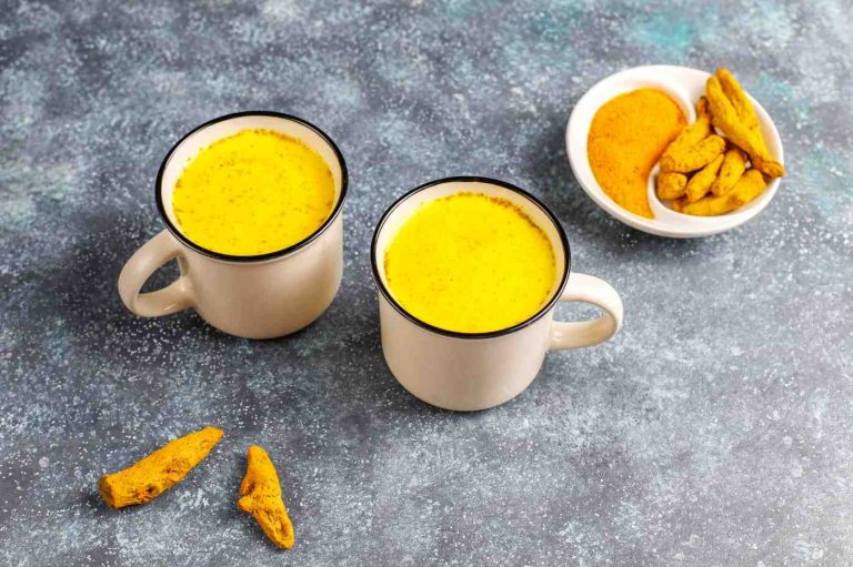 Two mugs of golden turmeric milk on a table with turmeric root and powder.