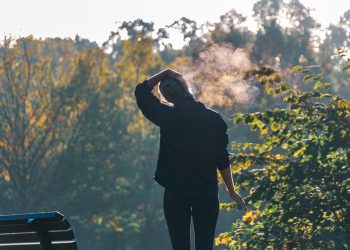 Person standing near bench in park, exhaling on a chilly morning.