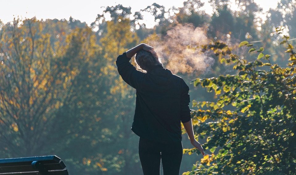Person standing near bench in park, exhaling on a chilly morning.