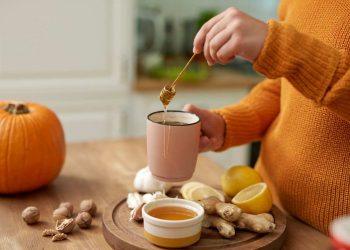 Pouring honey into a mug surrounded by ginger, lemons, and a pumpkin.
