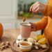 Pouring honey into a mug surrounded by ginger, lemons, and a pumpkin.