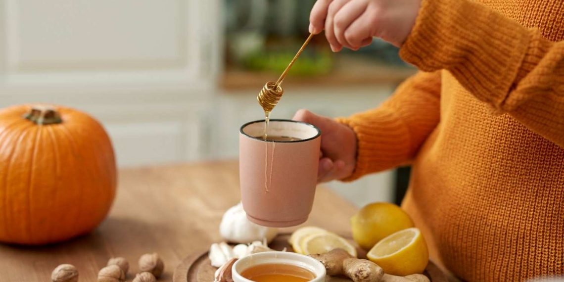 Pouring honey into tea with ginger, lemon, and garlic on table.