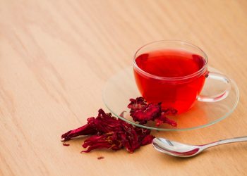 Glass cup of hibiscus tea with dried petals on wooden table.