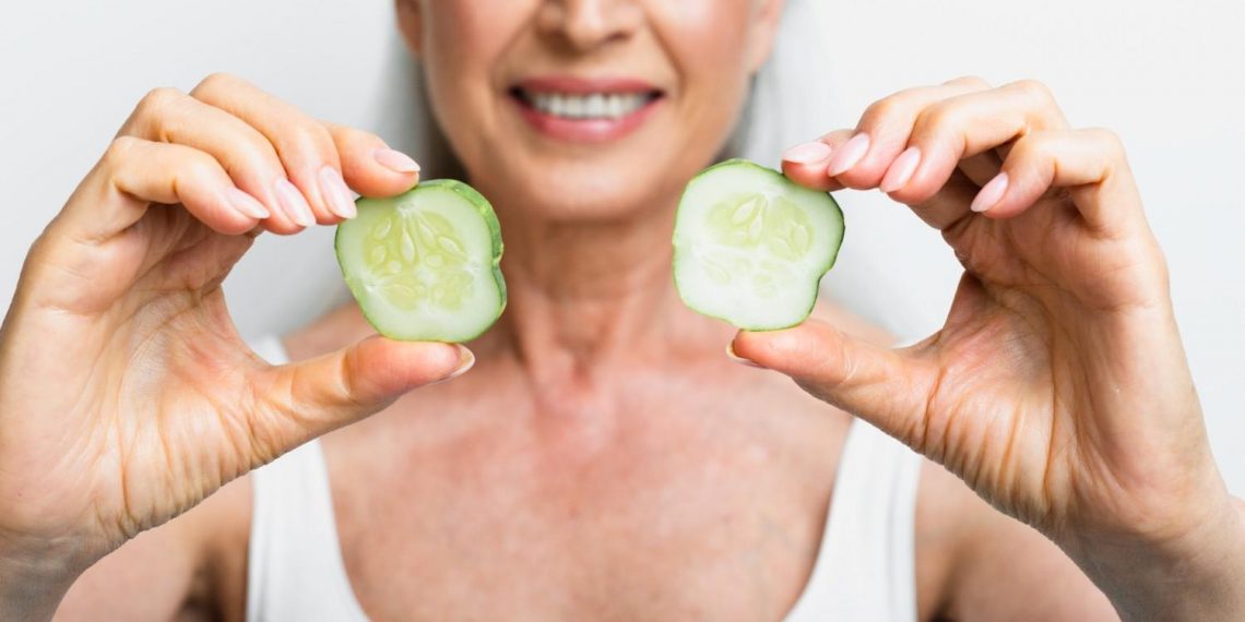 Senior woman smiling and holding cucumber slices.