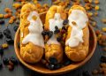 Cream eclairs topped with raisins on a wooden plate.