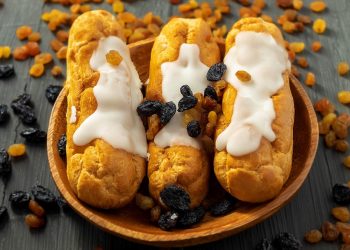 Cream eclairs topped with raisins on a wooden plate.
