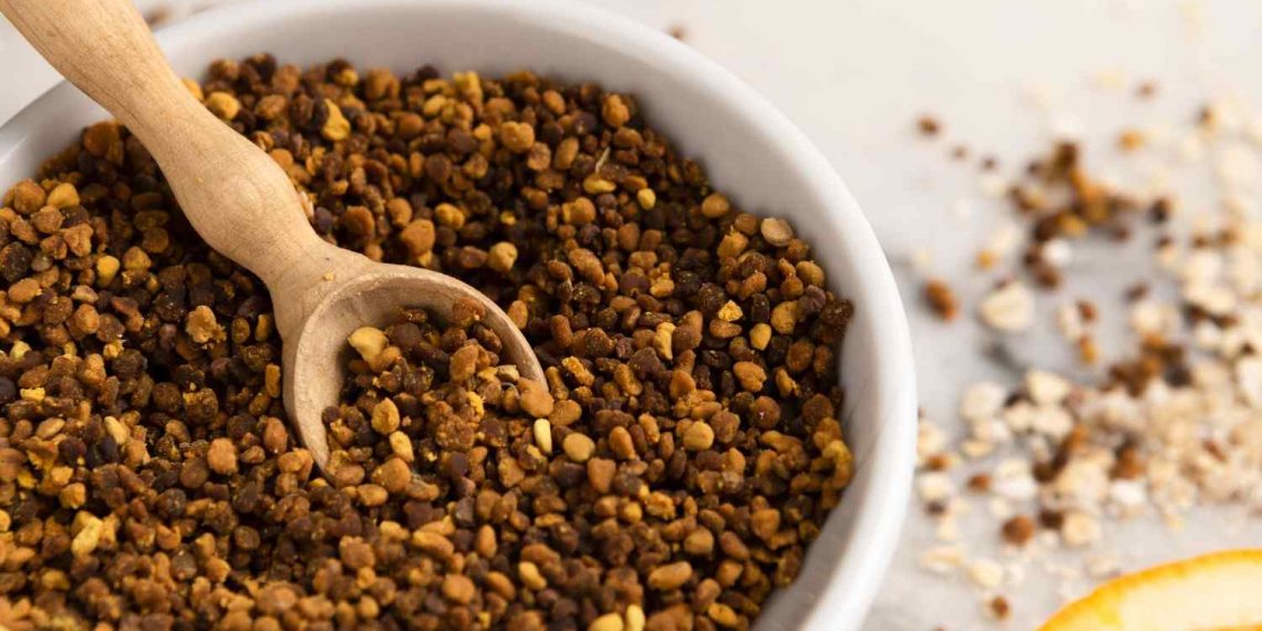 Bowl of bee pollen granules with wooden scoop on a table.