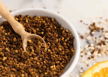 Bowl of bee pollen granules with wooden scoop on a table.