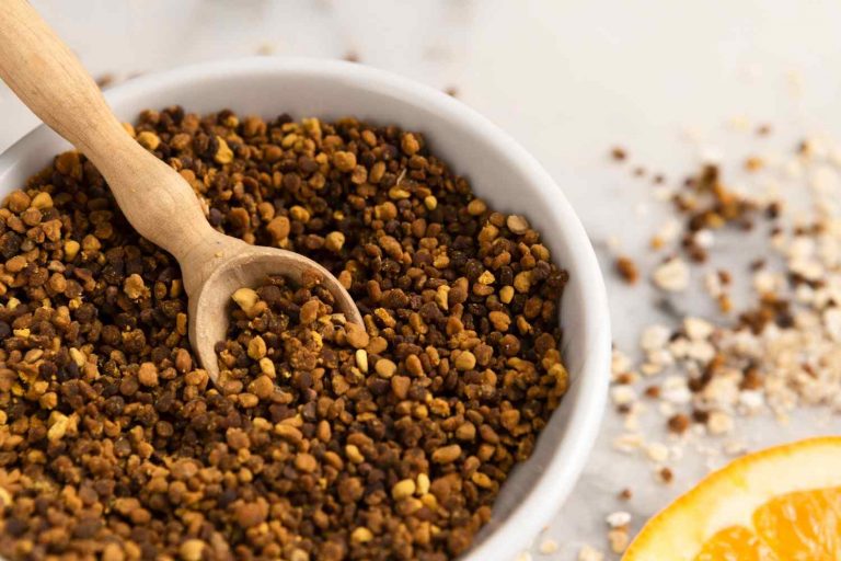 Bowl of bee pollen granules with wooden scoop on a table.