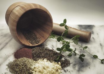 Wooden mortar with pestle, herbs, and spices on a marble surface.