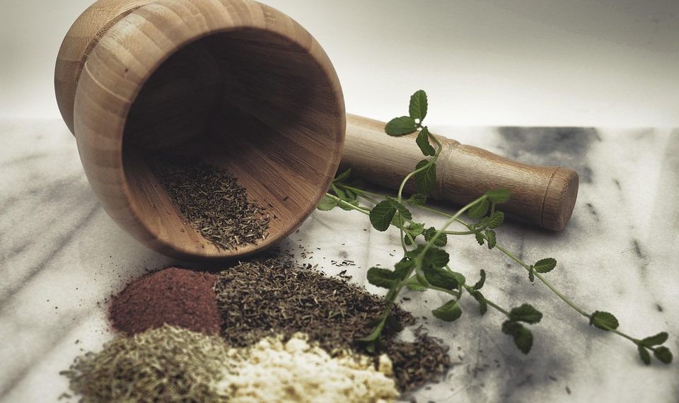 Wooden mortar with pestle, herbs, and spices on a marble surface.