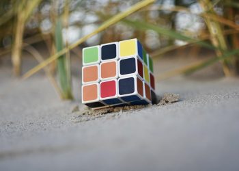 Rubik's Cube partially buried in sand at a beach, with strands of grass nearby.