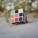 Rubik's Cube partially buried in sand at a beach, with strands of grass nearby.