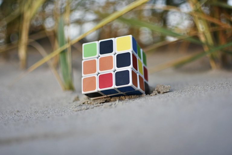 Rubik's Cube partially buried in sand at a beach, with strands of grass nearby.