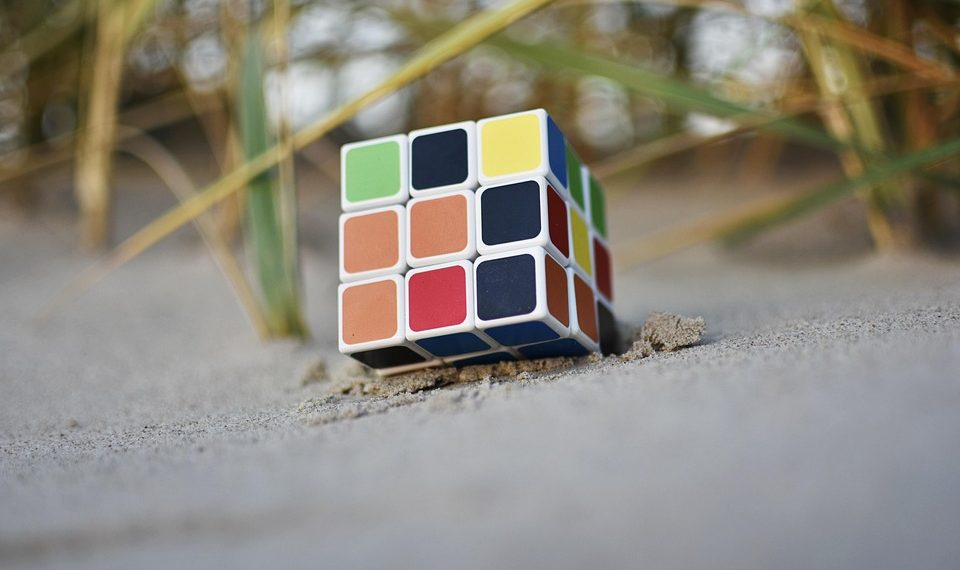Rubik's Cube partially buried in sand at a beach, with strands of grass nearby.