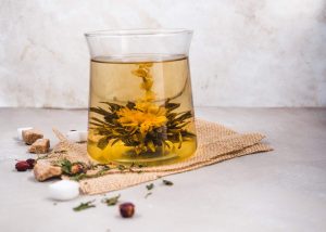 Blooming tea flower in a glass with sugar cubes and herbs scattered around.
