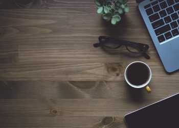 Laptop, coffee cup, glasses, plant on wooden desk.