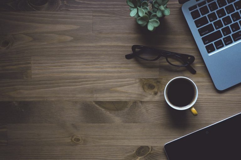 Laptop, coffee cup, glasses, plant on wooden desk.