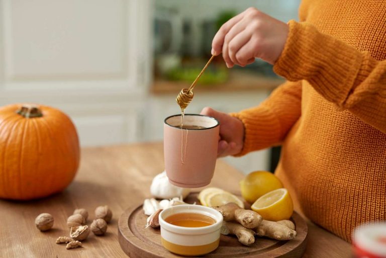 Pouring honey into a cup with lemon, ginger, and garlic on the table.