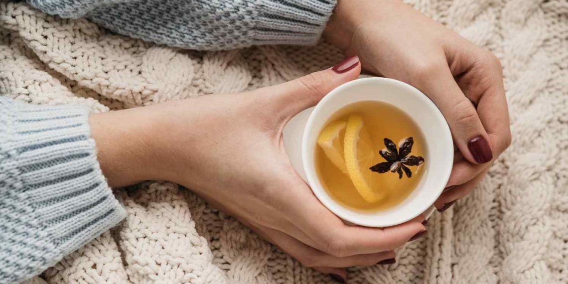 Hands holding a cup of warm tea with lemon and spice on a cozy knit blanket.