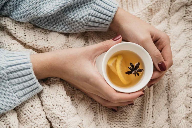 Hands holding a cup of warm tea with lemon and spice on a cozy knit blanket.