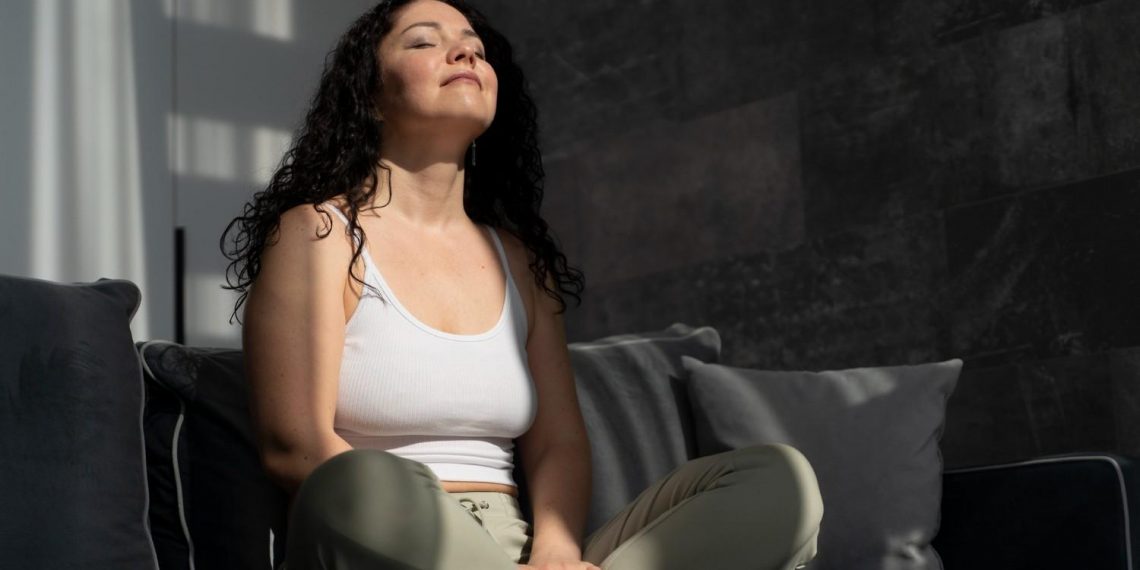 Woman meditating on couch, eyes closed, enjoying sunbeam.